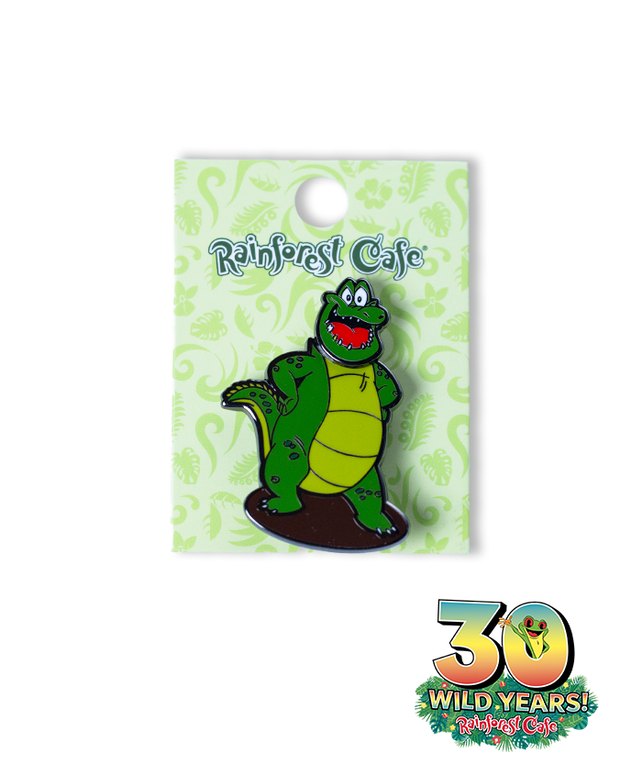 A collectible pin from Rainforest Cafe, featuring a cheerful green cartoon crocodile with a yellow belly. The crocodile stands upright and smiles broadly. It’s mounted on a light green decorative card adorned with darker green swirls and leaf patterns, with ‘Rainforest Cafe’ written in dark blue at the top. The card also celebrates the cafe’s 30th anniversary with ‘30 WILD YEARS!’ and a tree frog coming out the zero.