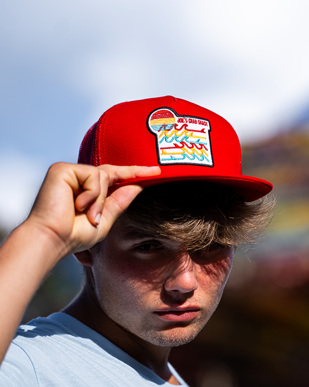 close up of male model wearing Red cap with red mesh back that has Joe's Crab Shack patch with red, yellow, and blue waves.
