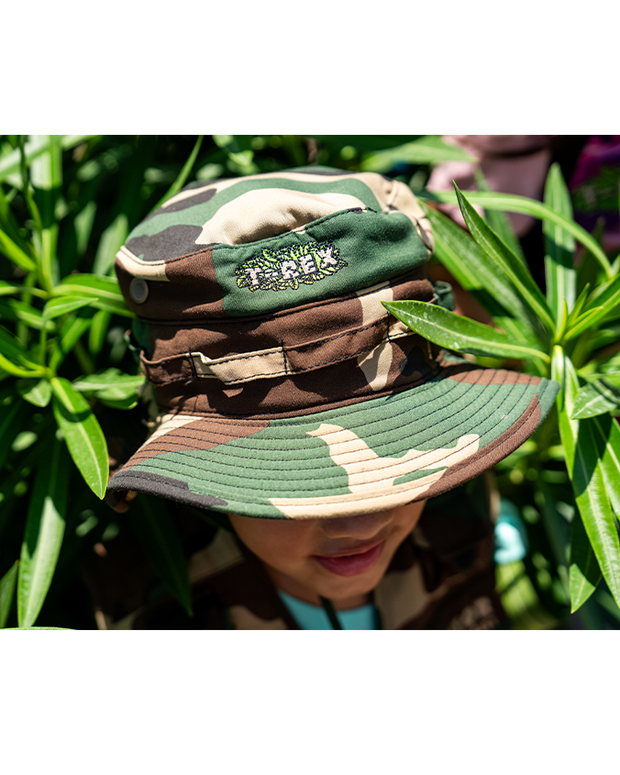 little boy standing in greenery wearing green camo hat.