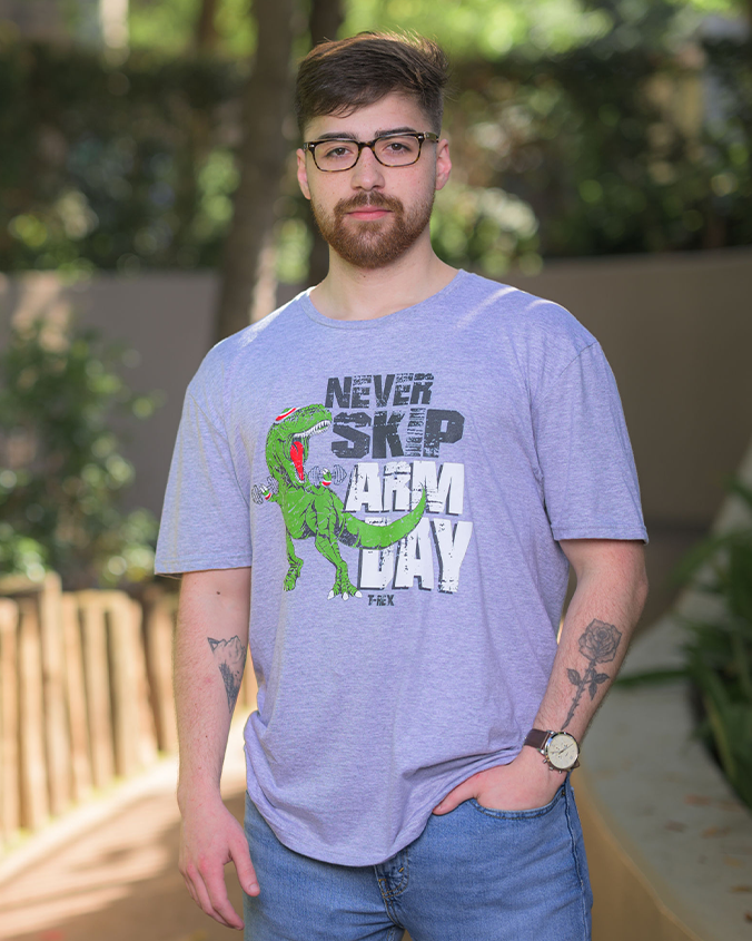A person wearing a grey t-shirt with a graphic of a green dinosaur and the text ‘NEVER SKIP ARM DAY’ printed on it, standing outdoors. He is wearing denim jeans and has one hand in his front pocket.