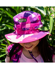 little girl wearing ranger hat, surrounded by greenery. close up of Pink camouflage safari hat with adjustable chin strap and T-Rex cafe logo embroidered in green. 