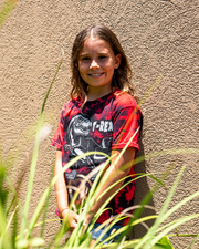 little girl leaning against wall wearing  Red and black tie-dye t-shirt with a T-Rex on center. Word "T-Rex" on top left side of t-rex image.