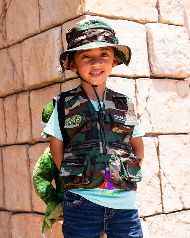 little boy standing in front of stone wall wearing matching green, camo vest and hat with a light blue shirt under vest and denim pants.