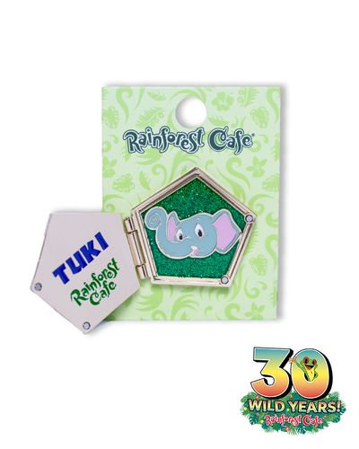 A collectible pin from Rainforest Cafe featuring Tuki, a cute grey elephant character, on a glittery green background, celebrating ‘30 Wild Years’ of the cafe, attached to a card with ornate patterns and the cafe’s logo.