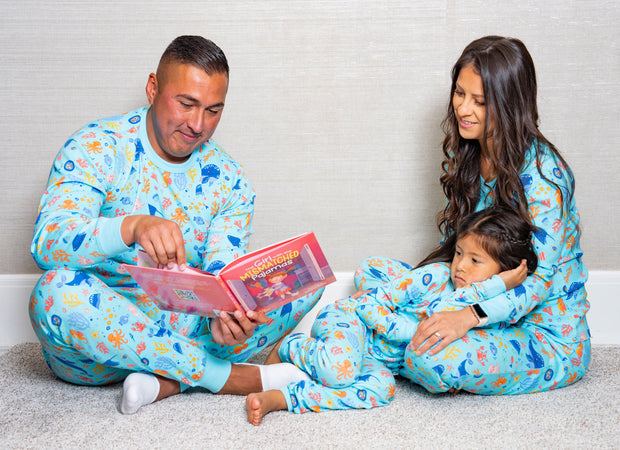 Mother and father reading a book to their daughter, as all of them are wearing the Under the Sea PJ sets