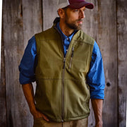 Man wearing olive vest, blue button down, brown jeans, and red trucker hat in front of rustic background.