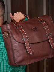 Man wearing green plaid long sleeve with bag over his shoulder by the handle in front of brown wood background.