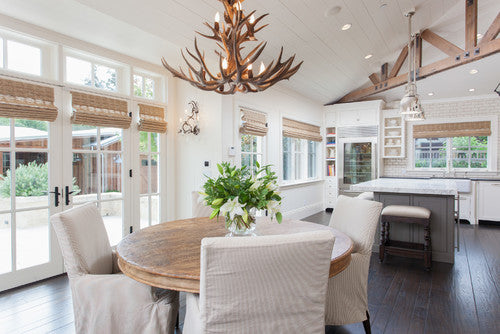 Elegant modern house with white and wood aesthetic in dining area that has antler chandelier above circular kitchen table. 