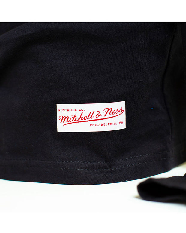 Close-up of the botom hem of the tee with Mitchel & Ness label. The label is white with red letters on it. The wording says: "Nostalgia Co. Mitchel i& Ness Philadelphia, PA."