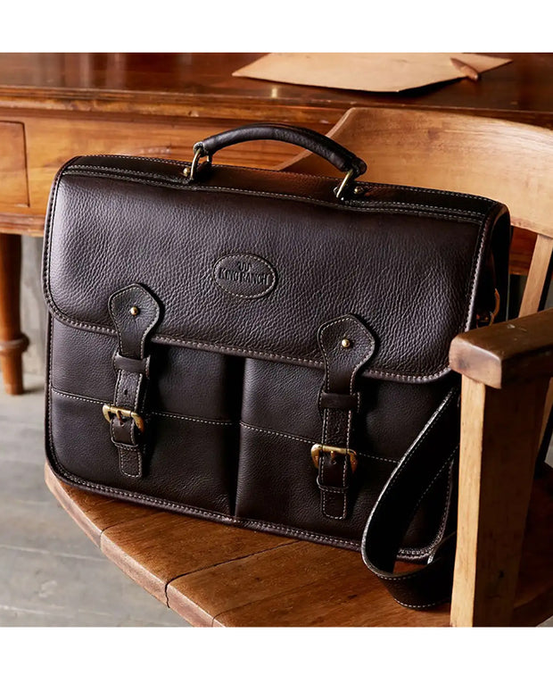 Dark brown briefcase features a front flap with two straps and buckle closures, a top handle, and an embossed King Ranch logo in the center. There is also an adjustable shoulder strap attached to the sides of the briefcase. Briefcase sitting on top a wooden chair.