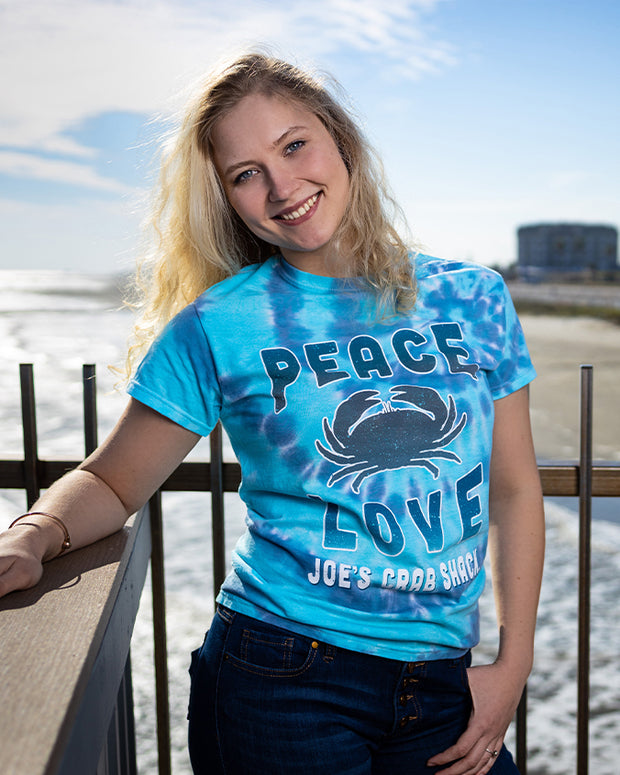 Blonde woman wearing Peace, Love & Crabs tee on pier in front of ocean in background.