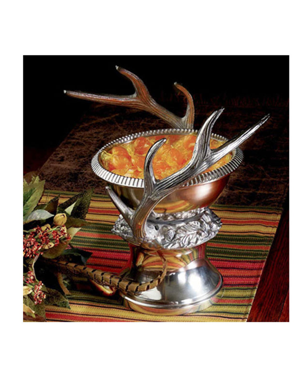 King Ranch Celebration bowl made of solid pewter with a mirror finishantlers on both sides, pictured with fruit inside, placed on table with flowers next to it.