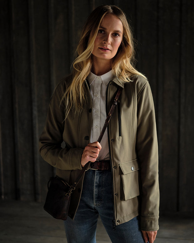 Blonde woman wearing leather tooled bag, white shirt, green jacket, and blue jeans in front of rustic background.