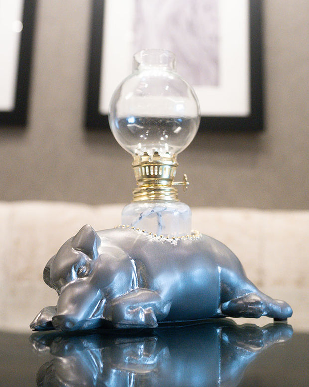 Silver pig laying down with oil lamp coming from its side placed on dining table in front of artwork wall.
