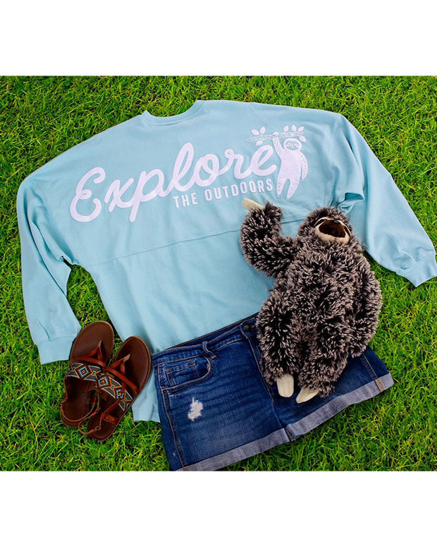 Light blue raglan with "Explore the Outdoors" written in white with a sloth decal laying in grass with shorts, sandals, and sloth plush.