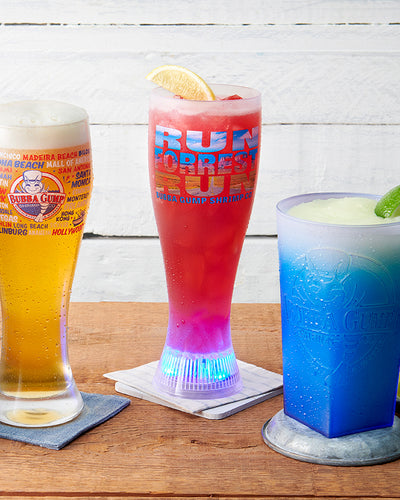 Three cups with refreshers in them. On the left, a Bubba Gump logo with store locations is printed on a pilsner glass. In the center, the Run Forrest Run Projector Glass has the image of the famous Highway 163 Scenic Drive within the words. On the right side is a Bubba Gump Logo Blue Shaker glass.