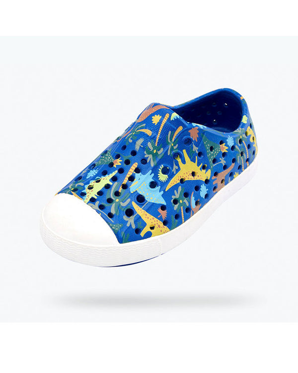 A single blue slip-on shoe with ventilation holes, a white toe cap, decorated with a colorful pattern of dinosaurs and plants in yellow, green, blue, and brown. 