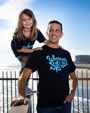 Man and little girls on a pier with beach in the background. The little girl is standing on wood rail, leaning in to the man. He is wearing the "Trouble Finds Me" tee and she is wearing matching shirt that just reads "trouble" in shark silhouette. 