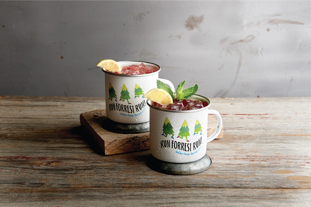 Two white tin mugs on wood table. Both are on metal coasters, and one topped on a wood raiser. Mugs have three pine trees running. Under them reads "RUN FORRESRT RUN!" and "Bubba Gump Shrimp Co." on bottom. Both drinks contains  red drinks with ice, topped with a lemon slice and one with mint leaves. 
