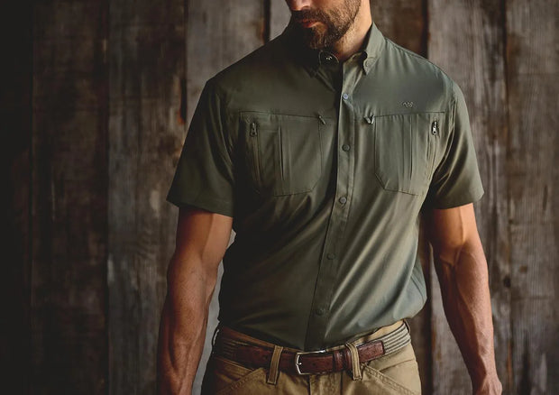 Bearded man wearing olive green short sleeve collared shirt tucked into belted pants in front of rustic background.