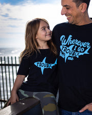 A man and little girl with the beach in their background. Little girl is sitting on a wood rail and is smiling, looking at the man. She is wearing the Trouble tee. He is wearing matching tee and that reads in blue letters "wherever I go trouble finds me".